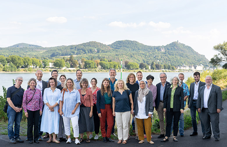 The participants of the 4th Round Table are standing together outside for a group photo, in the background there is the Rhine and parts of the Siebengebirge. All participants look very cheerful and a pleasant mood is noticeable.